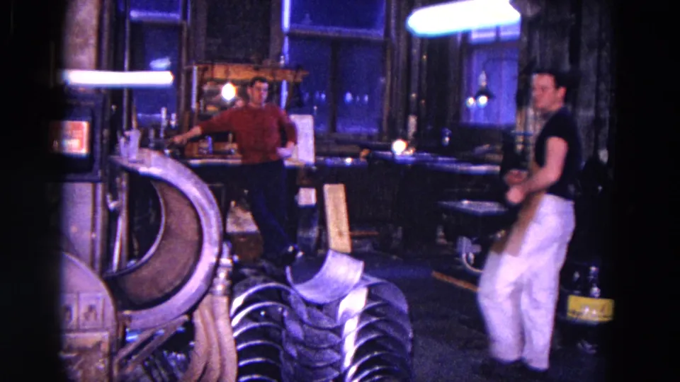 Two adult men appear in the 1967 Trenton shop interior, with one at the bench and another moving into frame in white pants.