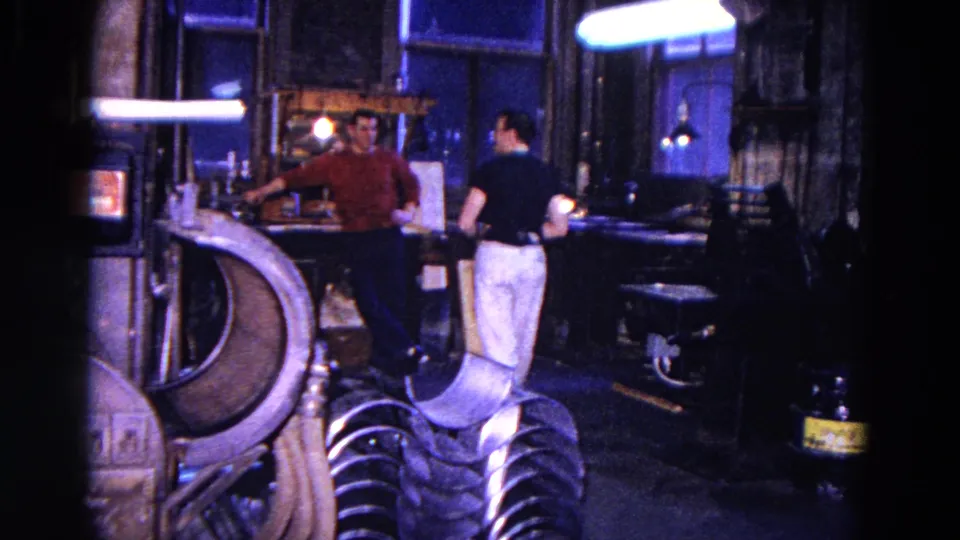 In the 1967 Trenton mechanic shop, two adult men stand near the bench as if taking a brief pause from work.