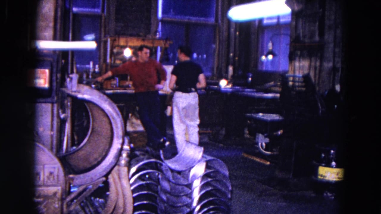 Two men stand and talk near machinery inside a dim workshop.