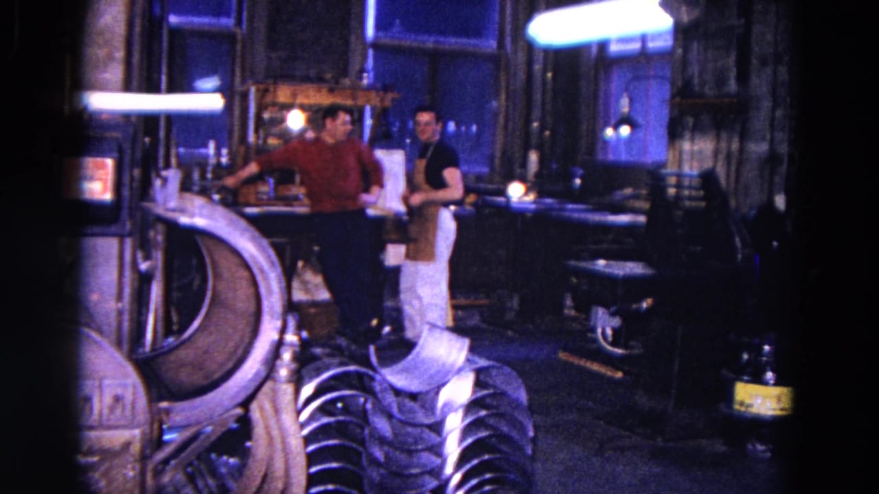Two men face each other near a bench with machinery around them.