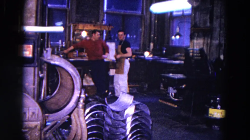 Two men appear to converse in a blue-lit 1967 Trenton workshop, surrounded by benches and metal equipment.