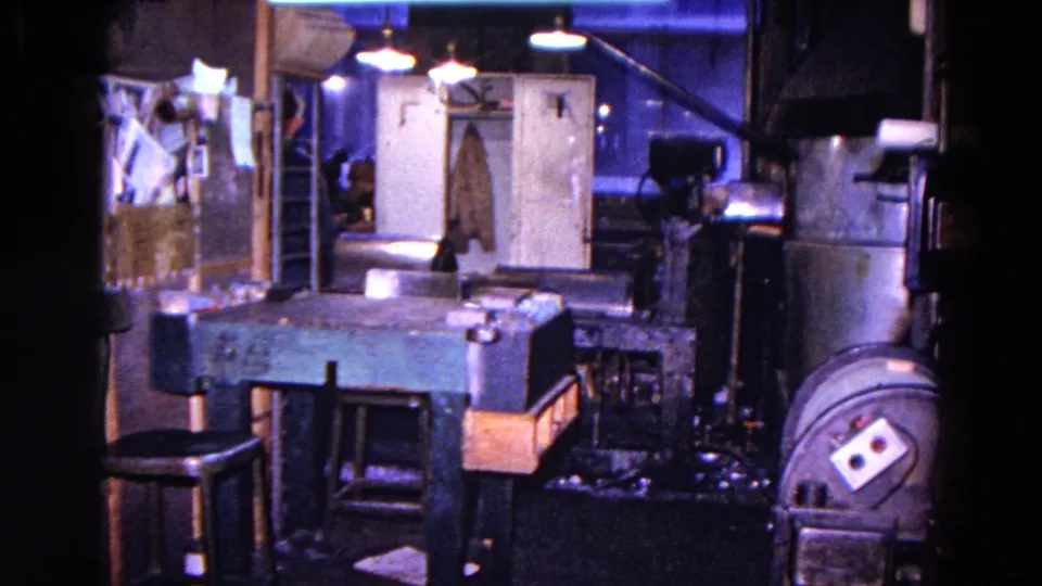 A dim 1967 Trenton workshop view shows tables, stools, a hanging coat, doors, and machinery in a crowded interior.