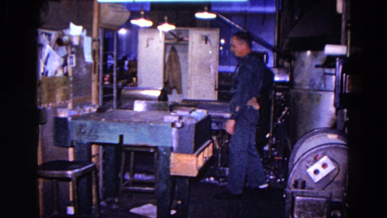 Worker in a dark uniform standing beside machinery and a workbench in a dim shop interior.