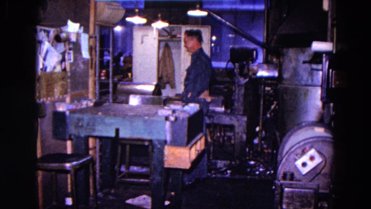 The worker appears beside large shop equipment, with the workbench, stools, lamps, wall notices, and doorway visible in the 1967 interior.