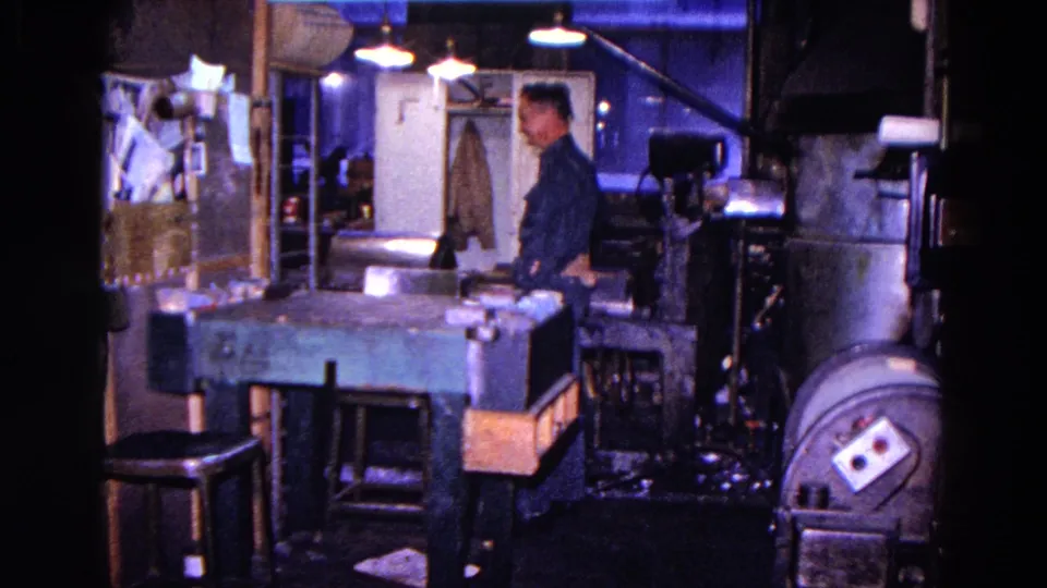 The worker appears beside large shop equipment, with the workbench, stools, lamps, wall notices, and doorway visible in the 1967 interior.