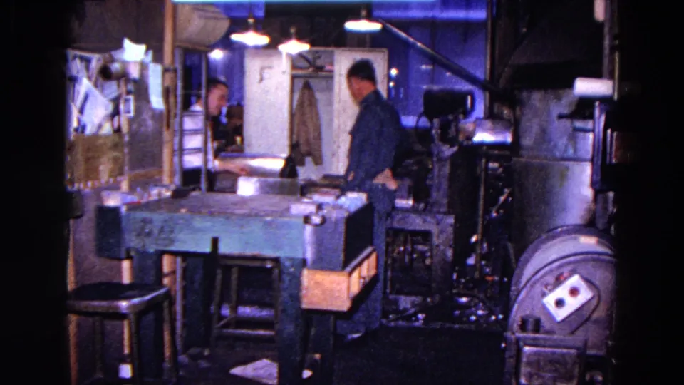 Two workers are visible in the Trenton machine-shop interior, one in a dark uniform and another partly seen near the back area.