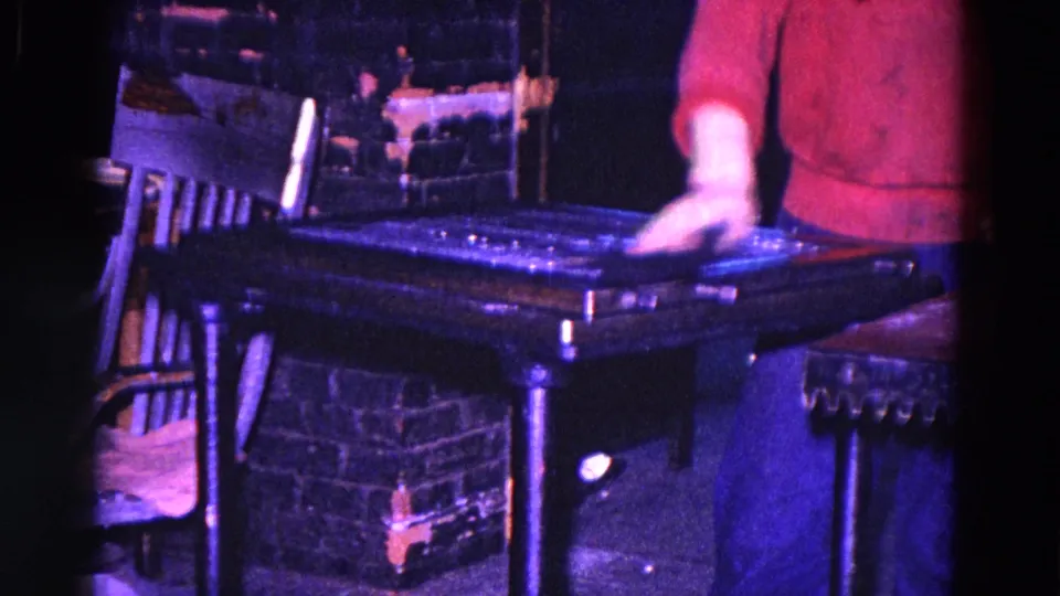 A man in a red shirt and blue pants wipes a dark metal table in the Trenton workshop, with a wooden chair and crates nearby.