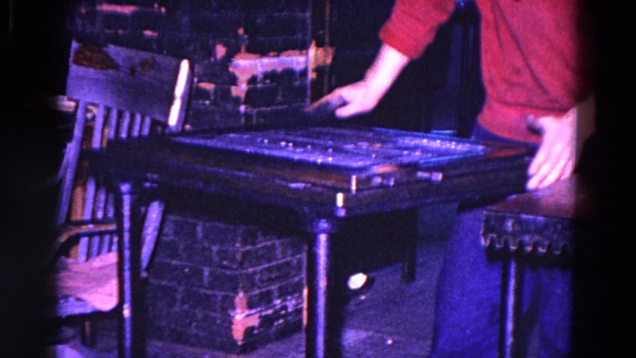 In 1967 Trenton, New Jersey footage, a red-sleeved person cleans a dark industrial machine beside a chair and brick wall.