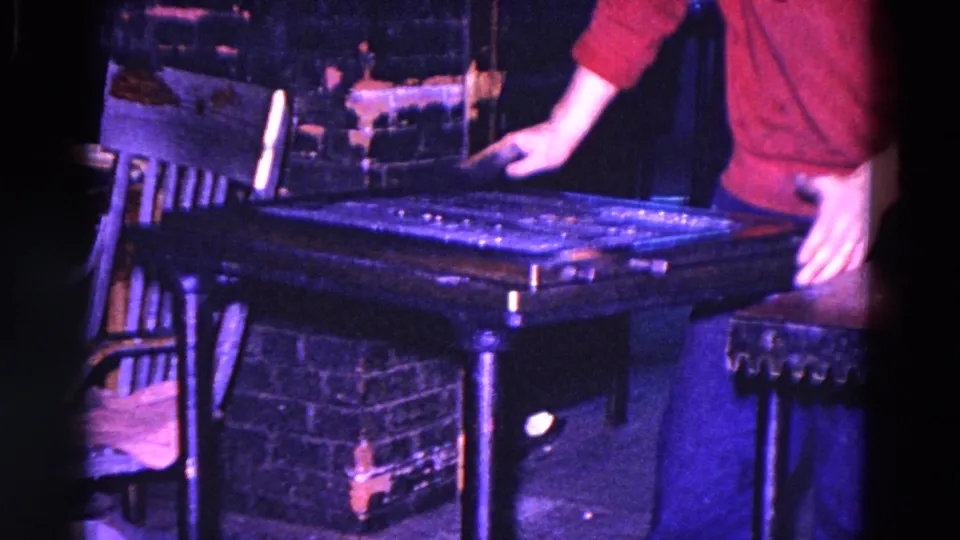 In 1967 Trenton, New Jersey footage, a red-sleeved person cleans a dark industrial machine beside a chair and brick wall.