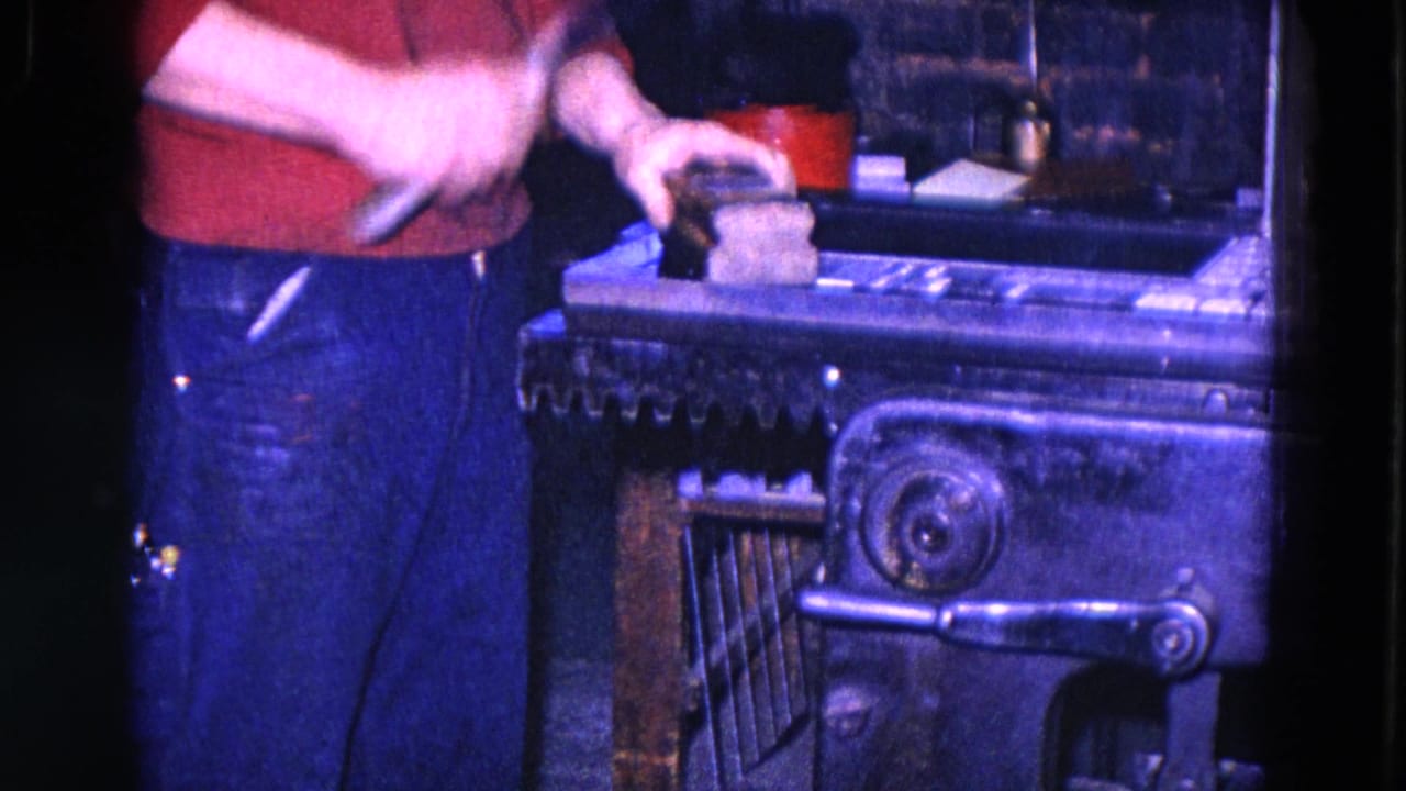 Worker in a red shirt holds a hammer near a metal block on forge equipment.