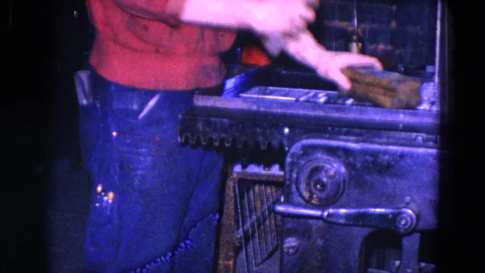 A close 8mm view shows the man in red and blue jeans reaching over the forge area while handling a rectangular metal block.
