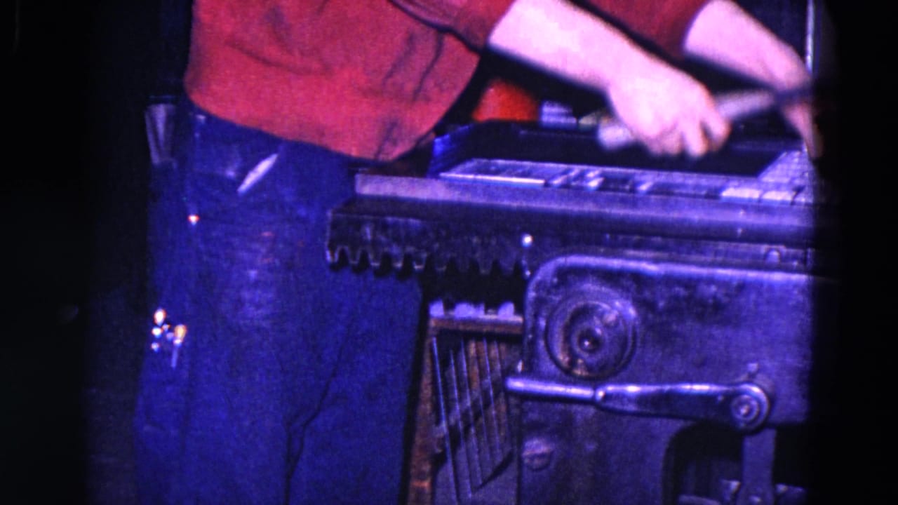 Worker in a red shirt reaches over a metal industrial press with levers and rollers visible.