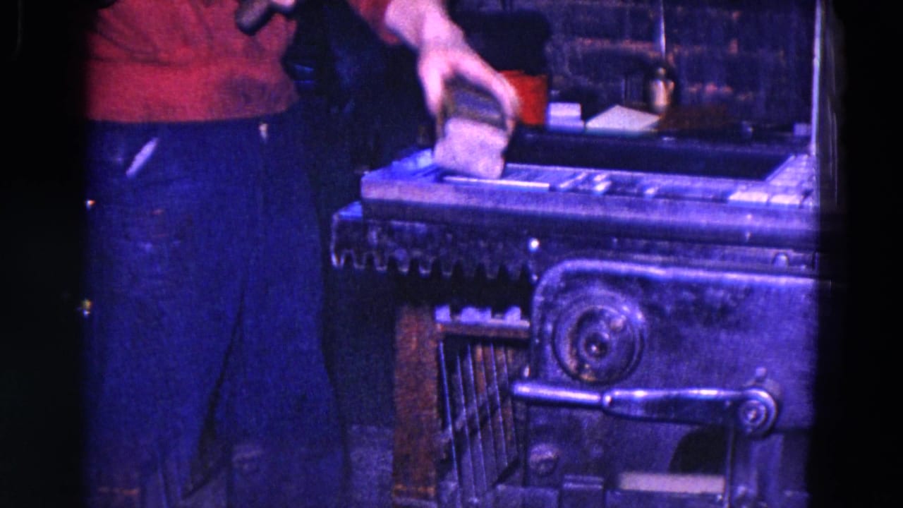 1967 Trenton 8mm frame with a worker's torso and hand holding material over an industrial printing press under blue-purple light.
