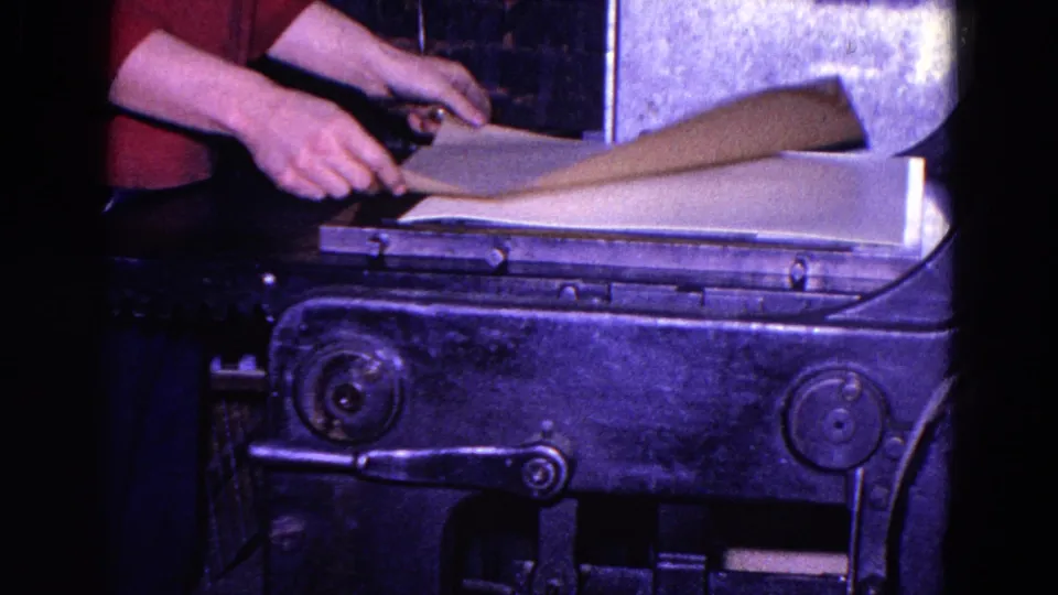 Hands lift and adjust a large sheet of paper on a worn industrial machine in this 1967 Trenton 8mm frame.