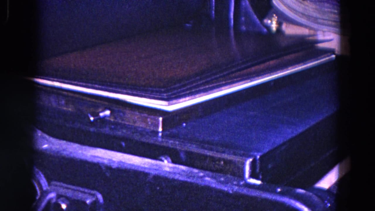 Close 8mm frame from Trenton, New Jersey, showing a dark book or cover moving through a press or conveyor area in a dim industrial setting.
