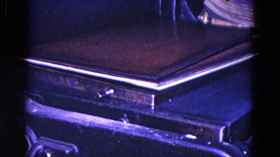 1967 Trenton close-up of book or cover material on a dark conveyor or press surface, with metal edges and strong blue-purple 8mm color shift.