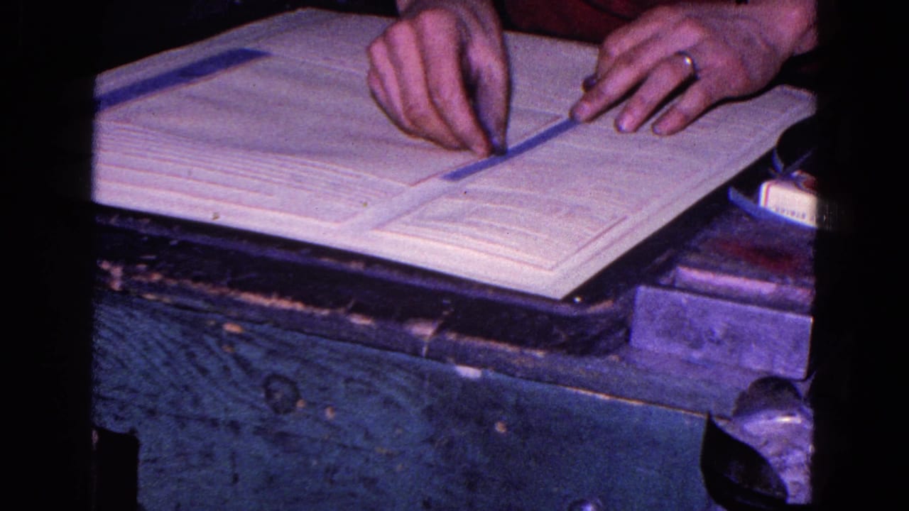 In 1967 Trenton home-movie footage, ringed hands align a narrow paper strip across a large document on a worn dark tabletop.