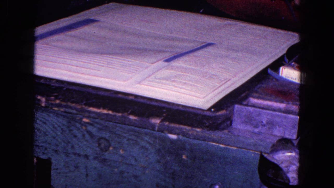 A close view shows the paper strip across the large document on a worn dark table, with purple-tinted 8mm color and soft focus.