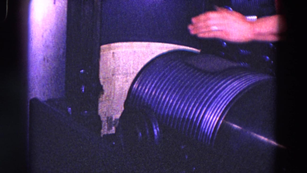 Close archival view of a hand working above a ribbed machine cylinder, with pipes and dark housing visible.