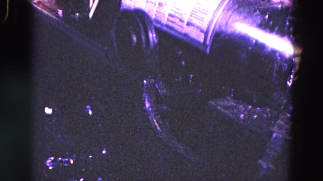 Close view of dark machinery and a shiny cylindrical metal part under purple-tinted light.