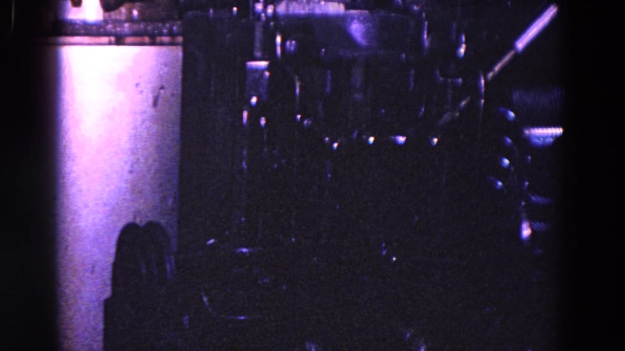 Dim factory machine with cylindrical equipment and a narrow bright tool or pipe at right.