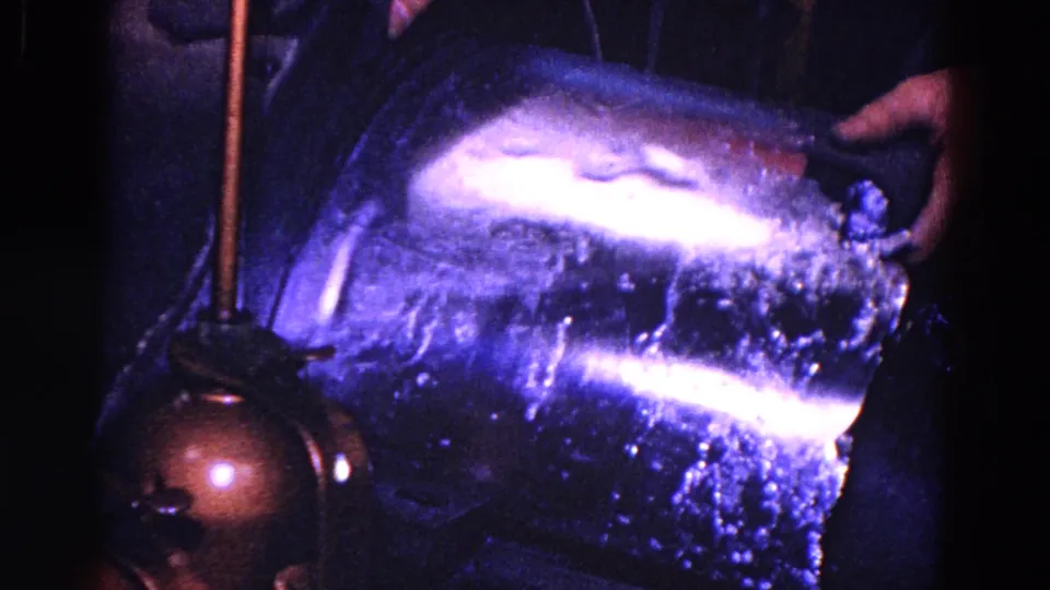 1967 Trenton 8mm close view of a hand handling a reflective cylindrical metal vessel beside factory machinery.