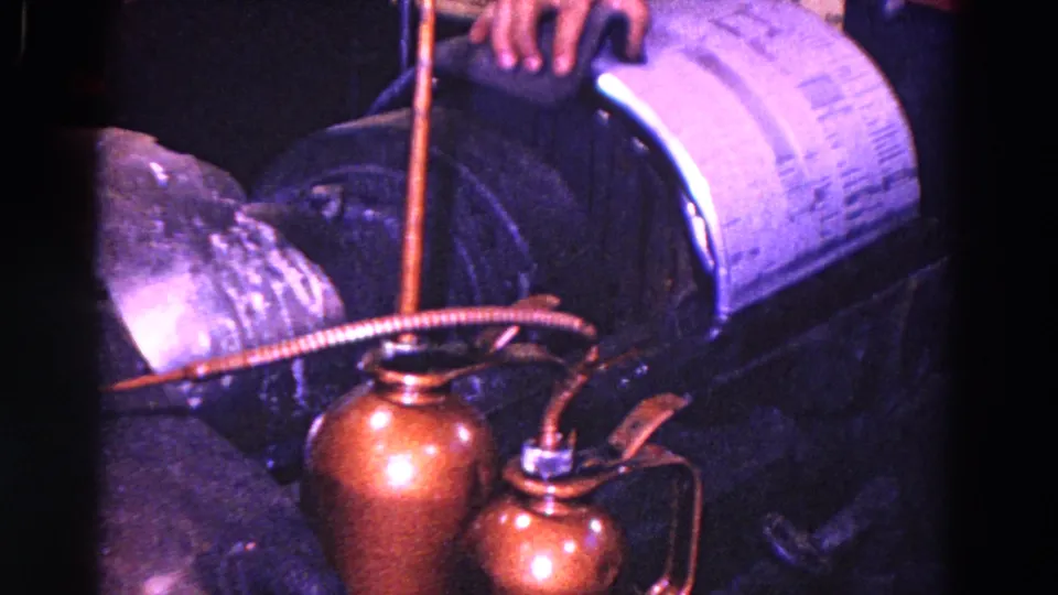 Trenton machine-shop frame with two copper oil cans, long spouts, hoses, a worker's hand, and heavy equipment in close view.