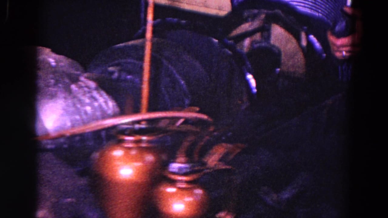 1967 Trenton industrial home-movie frame showing a tool working beside a corrugated metal tube, with reddish containers and heavy film shadow.