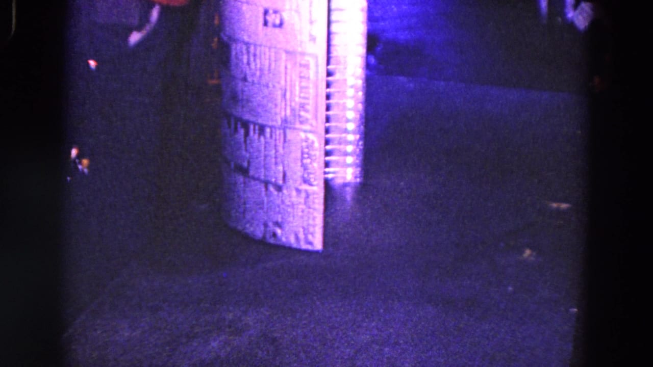 Corrugated metal tube stands on a dark surface with bright purple-blue light and heavy shadow.