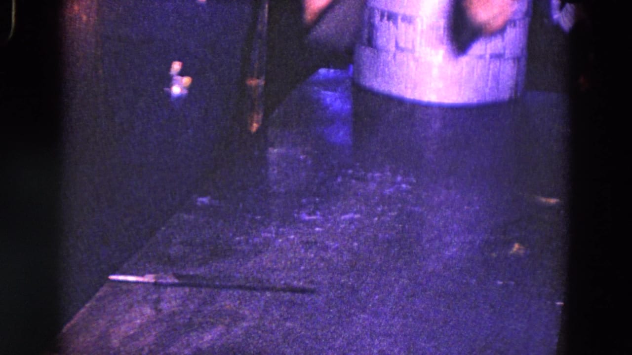 1967 Trenton home-movie frame with a cropped standing adult, dark floor area, and small objects visible in low purple-tinted light.