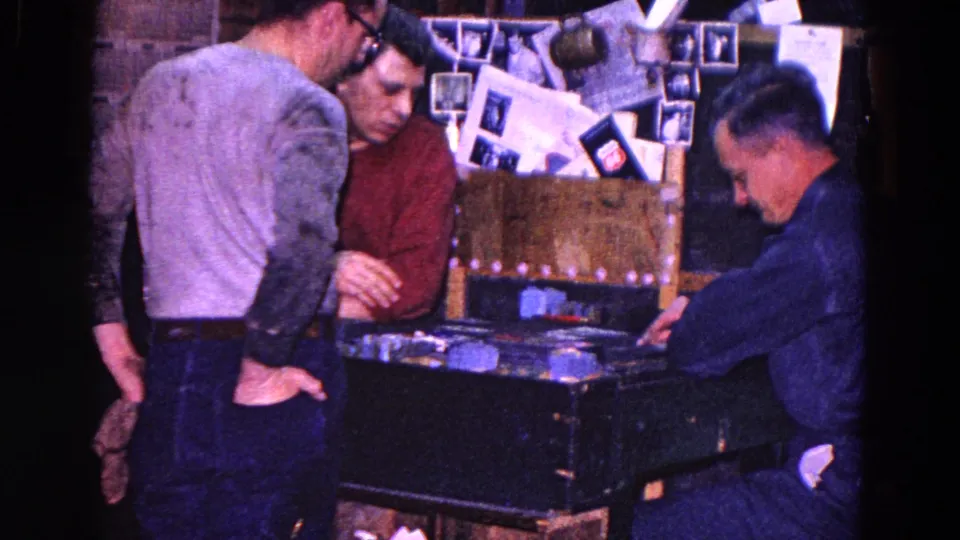 1967 Trenton 8mm frame showing adult men around a card table in a private interior with photographs and papers pinned behind them.