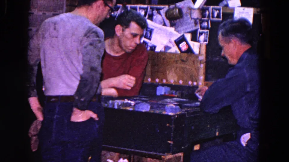 1967 Trenton home-movie frame of adult men focused on a tabletop card game in a dim private room with pinned wall material behind them.