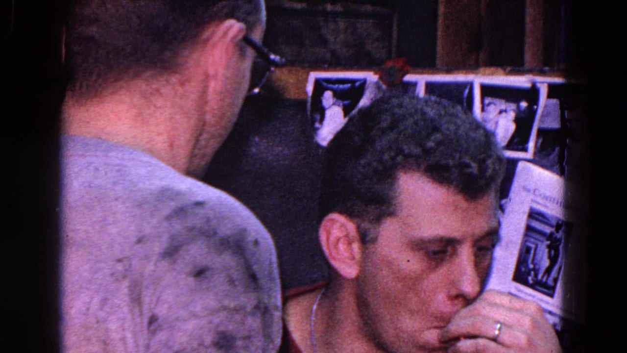 1967 Trenton home-movie frame showing adult men close to a tabletop game, with a man in glasses seen from behind and wall photos in the room.