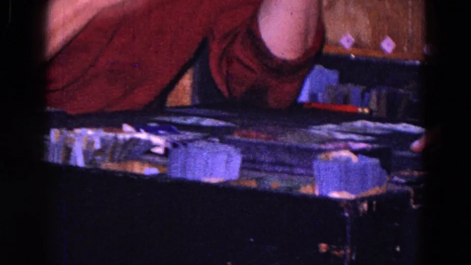 1967 Trenton home-movie frame showing a man leaning across a tabletop game, with stacked pieces and nearby adults partly visible.