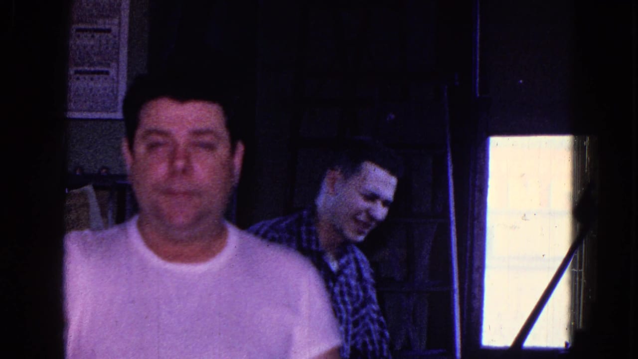 Inside the Trenton factory setting, a man in a white shirt faces the camera as a plaid-shirted coworker stands near a bright window.