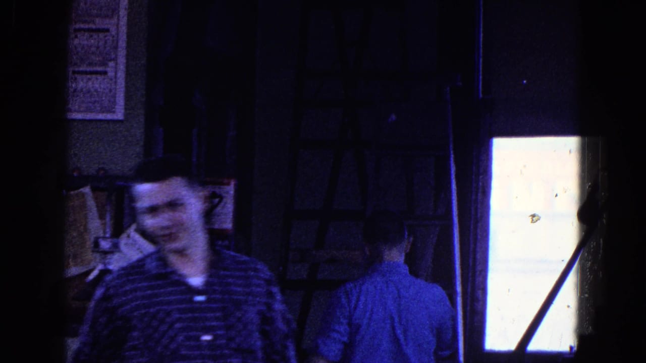 1967 Trenton 8mm frame showing two adult factory workers in a dark interior, one facing the camera and another turned away near equipment.