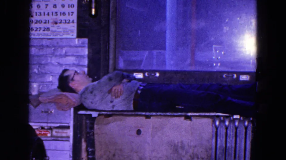 Adult man in glasses rests on a makeshift bed atop a metal bench in a 1967 Trenton industrial interior.