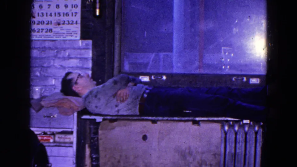 In the Trenton factory scene, a man lies on a bench with a pillow, calendar, brick wall, and radiator visible behind him.