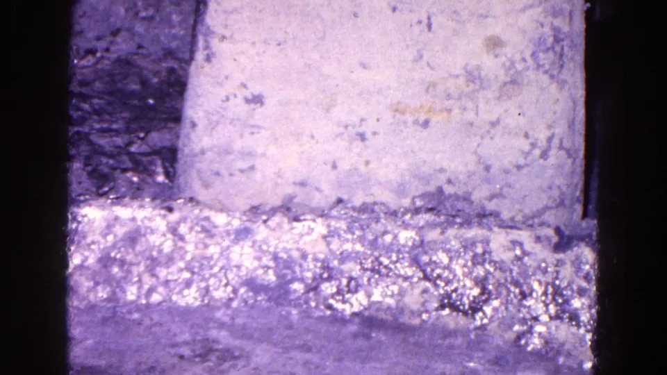 1967 Trenton 8mm frame of a textured pale block and shimmering liquid, filmed in tight close-up with dark vertical borders.