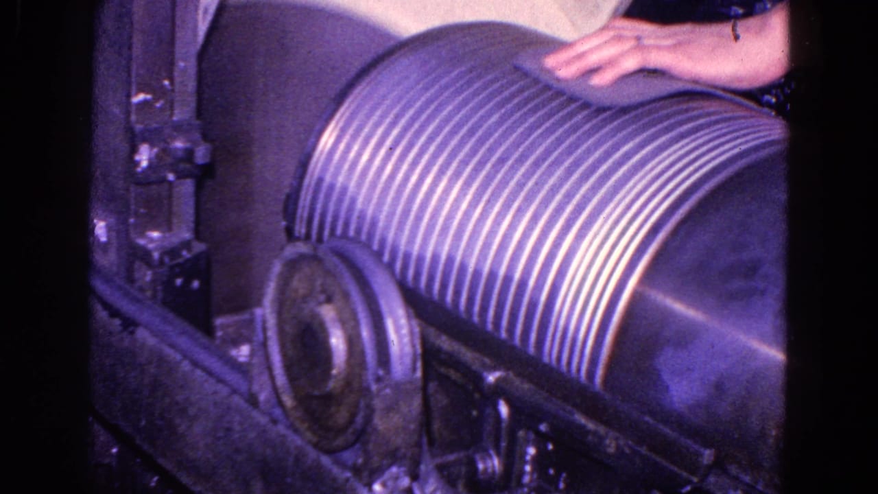 1967 Trenton, New Jersey 8mm frame showing a worker's hand above a ribbed cylindrical roller within dark industrial machinery.