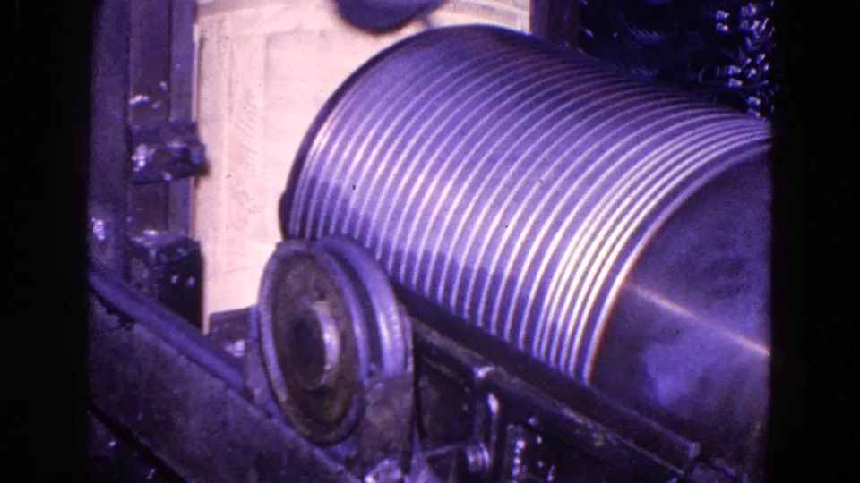 Close 8mm view of the ribbed cylindrical roller and machine housing, with a worker's light clothing partially visible above it.