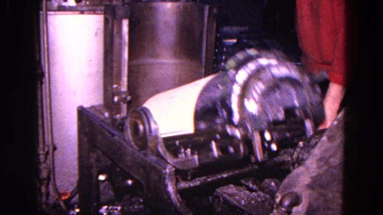 Worker's hands guide a metal canister on a dark industrial machine.