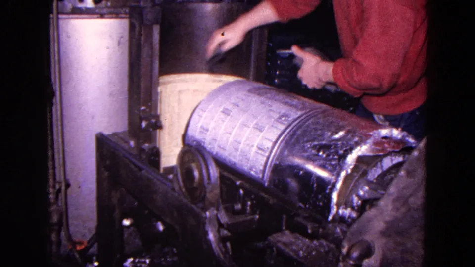 A worker in a red shirt leans over the machine while the large canister remains seated on the rollers.