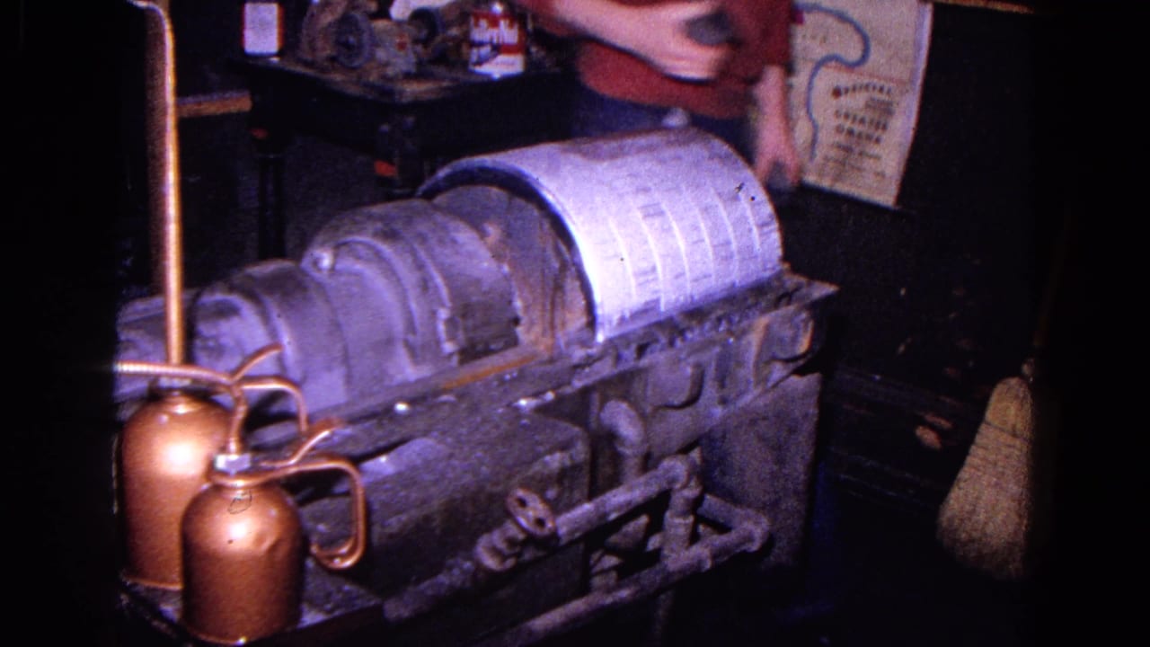 1967 Trenton home-movie frame showing a gray linen processing machine, copper oil cans, pipes, and a wall map inside a dim workshop.