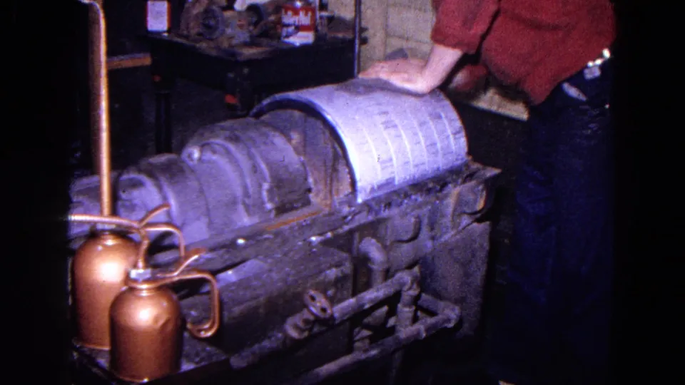 1967 Trenton home-movie frame showing a worker in a red sweater handling or steadying linen on a heavy processing machine.