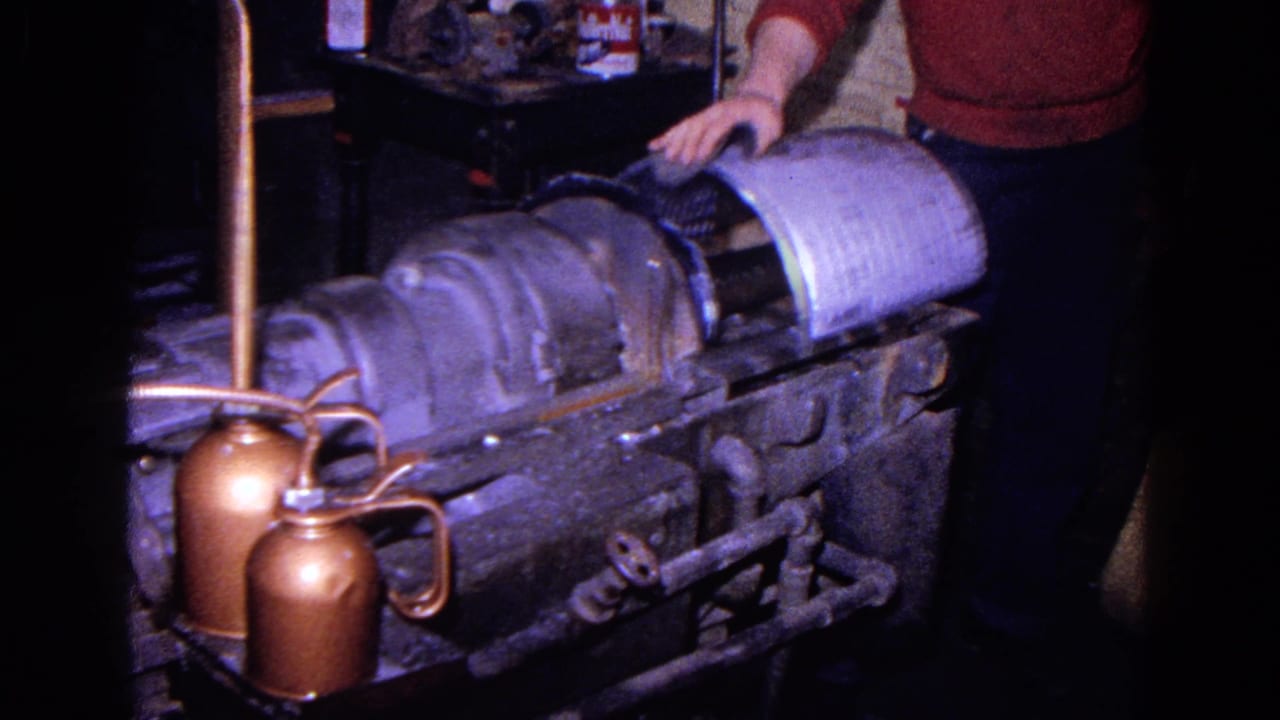 Worker stands beside a heavy machine with a metal cylinder and copper oil cans.