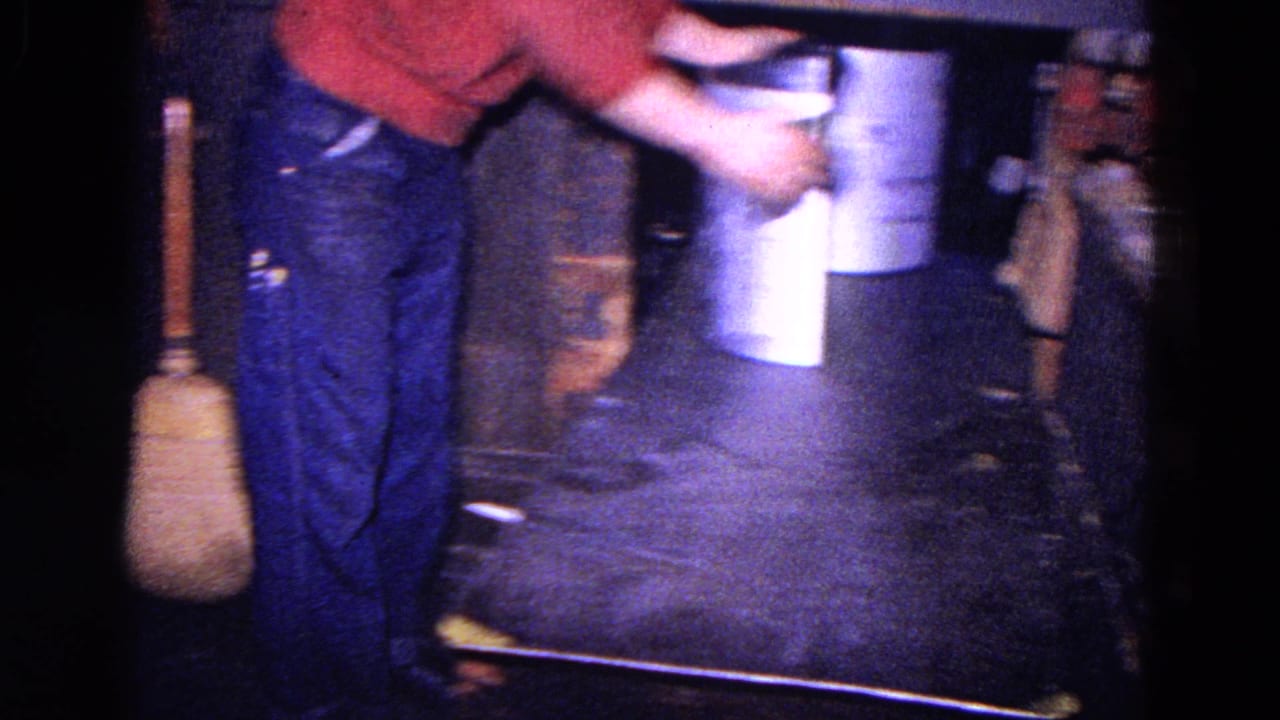 Worker carries a cylindrical metal piece near buckets and a broom in a dim workshop.