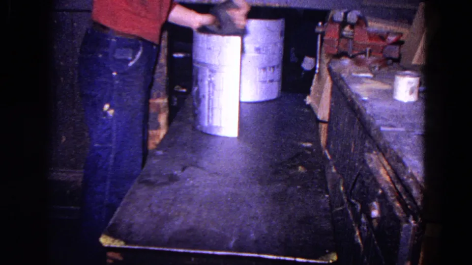 A worker in jeans holds a metal cylinder near a bench and vise inside the 1967 Trenton workshop scene.