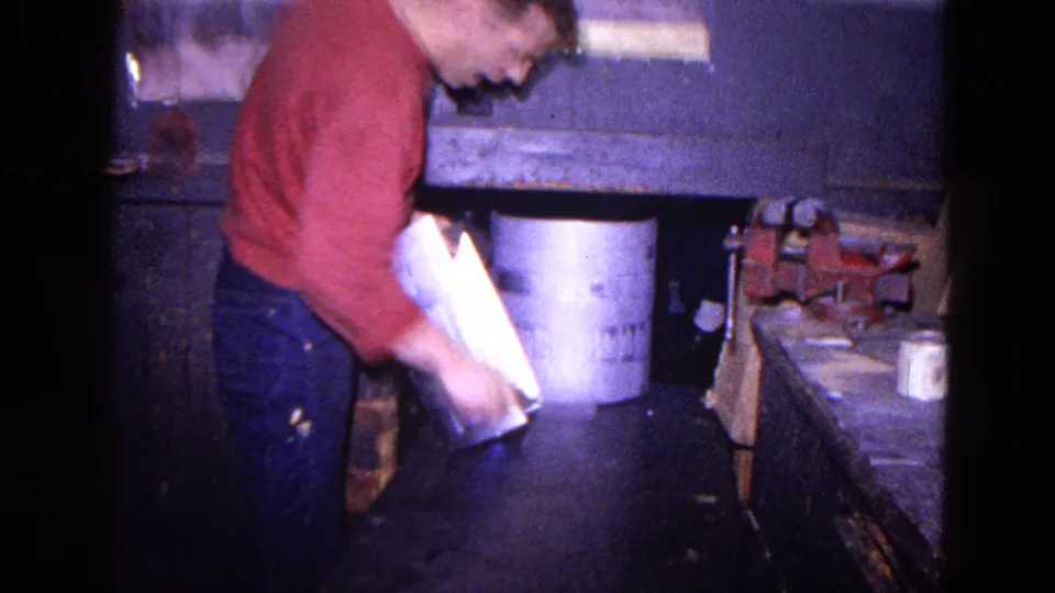 In the workshop interior, the worker bends while handling a cylindrical metal piece beside buckets, a bench, and a red vise.
