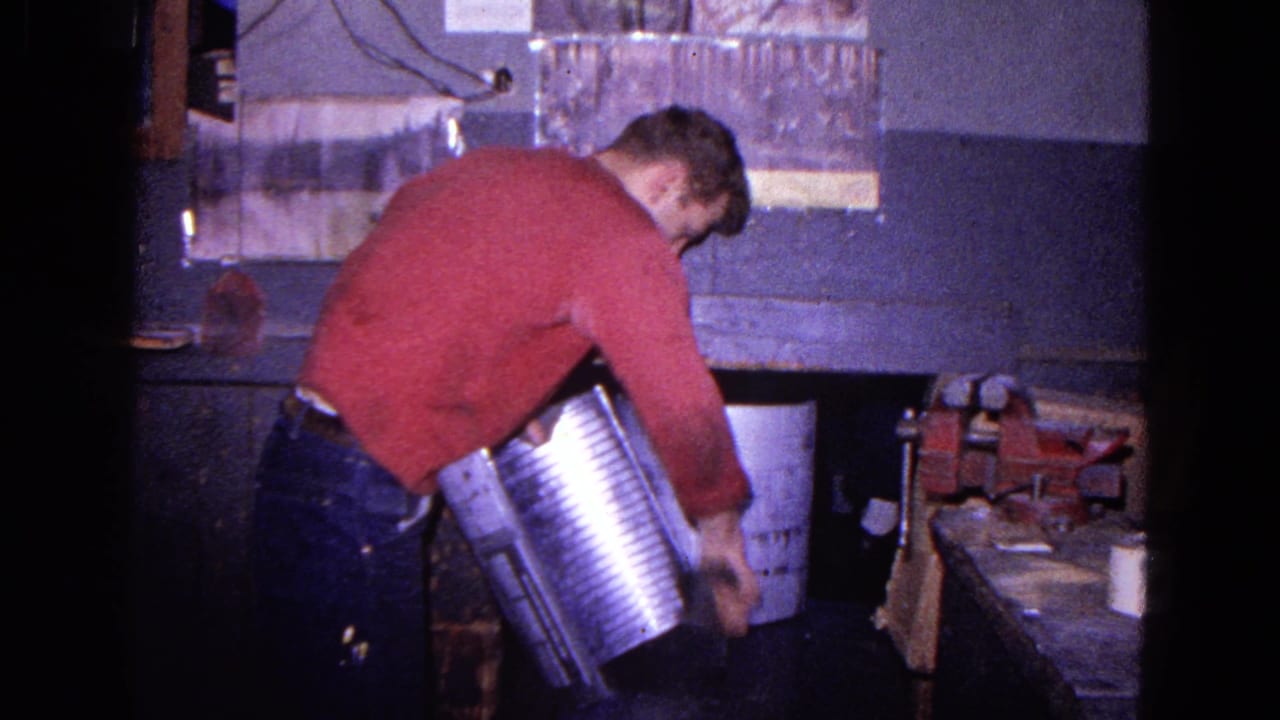 Man in a red sweater bends over a workbench while holding a large metal cylinder.